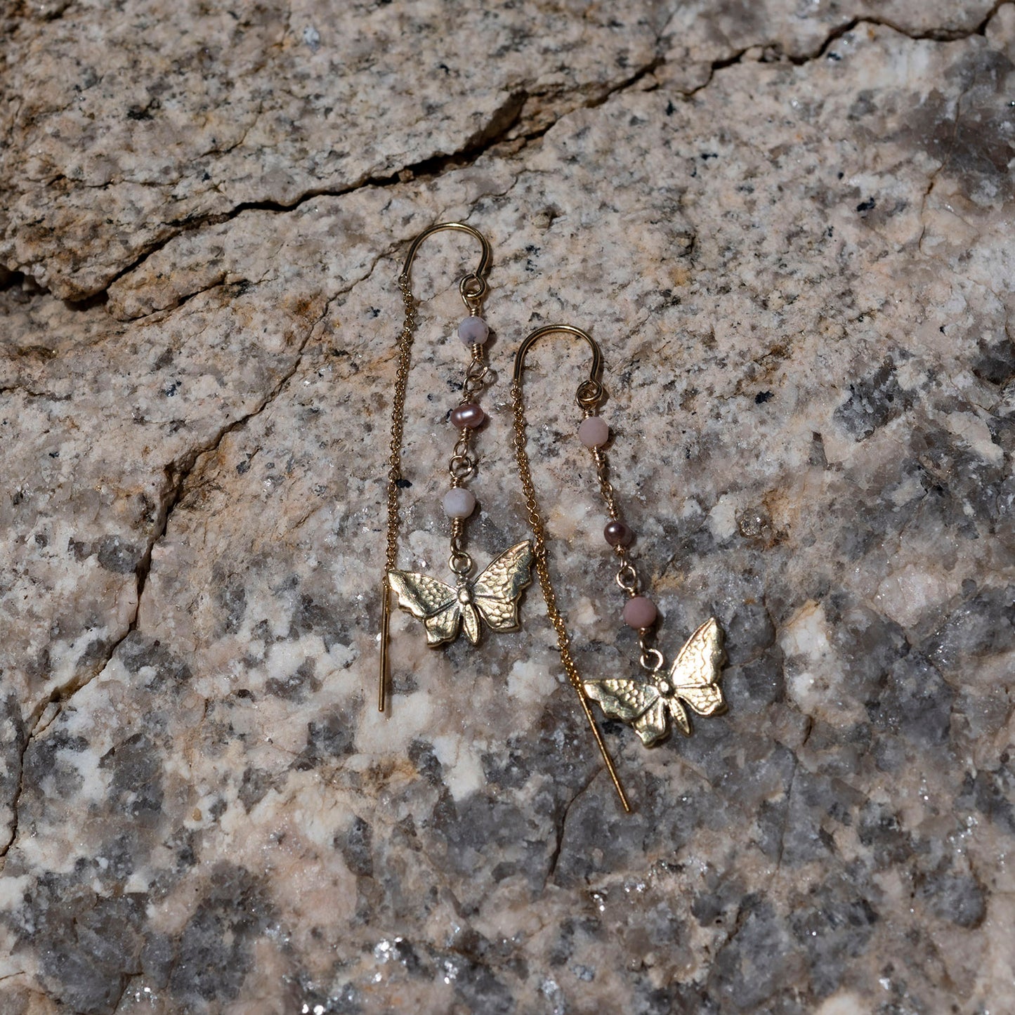 A pair of dangling earrings with gold-filled butterfly charms, opal and freshwater pearls beads, and a long drop chain, photographed on a textured stone surface.