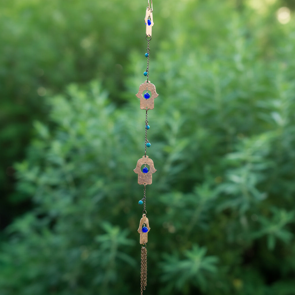 A hanging Hamsa wall hanging ornament with four copper hamsa charms, each featuring a Lapis blue evil eye gemstone, suspended from a chain with Turquoise beads, set against a blurred wooden fence and green plants.
