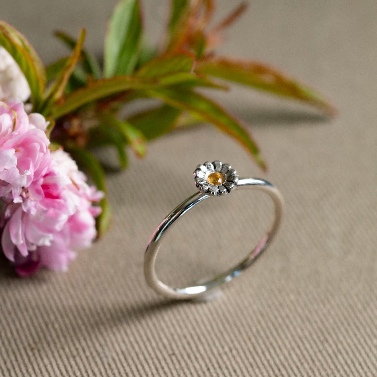 Silver daisy ring with a small yellow citrine on a beige surface with pink flowers and green leaves.
