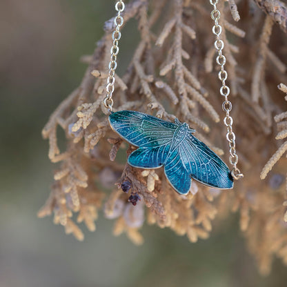 A silver-tone necklace with a detailed blue-green moth pendant, hanging against a soft-focus background of brown branches.