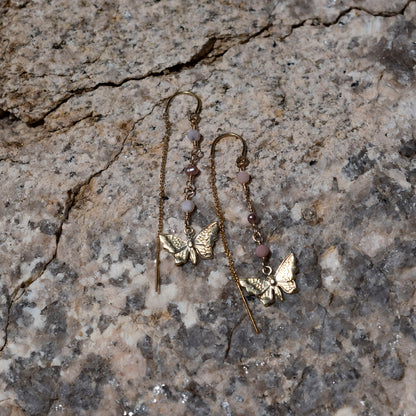A pair of dangling earrings with gold-filled butterfly charms, opal and freshwater pearls beads, and a long drop chain, photographed on a textured stone surface.