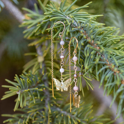 Gold-toned dangle earrings with a butterfly charm, pale pink beads, and small metallic charms, hanging from a pine branch outdoors.