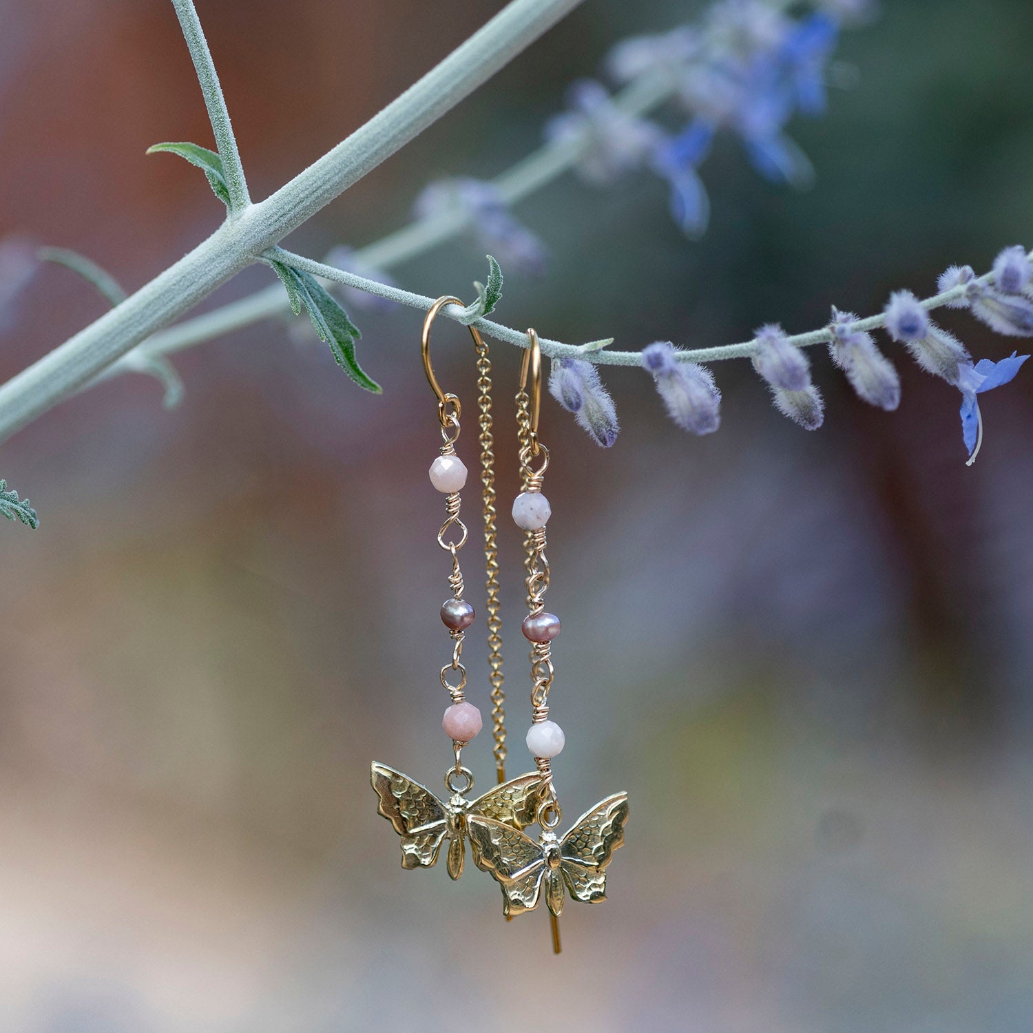 Gold-filled dangle earrings with butterfly charms and small pink and white beads, hanging from a plant stem with blurred lavender flowers in the background.