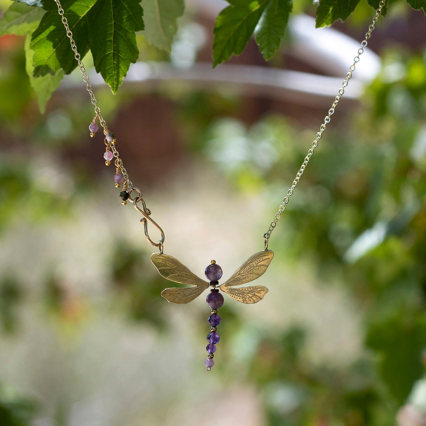 A delicate gold-toned chain necklace featuring a dragonfly pendant made of gold-toned metal with purple gemstone accents and a dangling purple bead drop, photographed outdoors against a blurred green leafy background.
