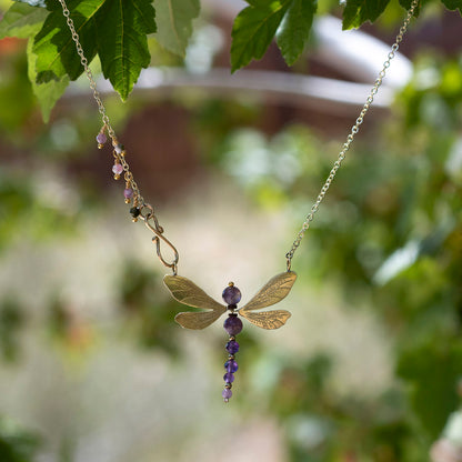 A delicate gold-toned chain necklace featuring a dragonfly pendant made of gold-toned metal with purple gemstone accents and a dangling purple bead drop, photographed outdoors against a blurred green leafy background.