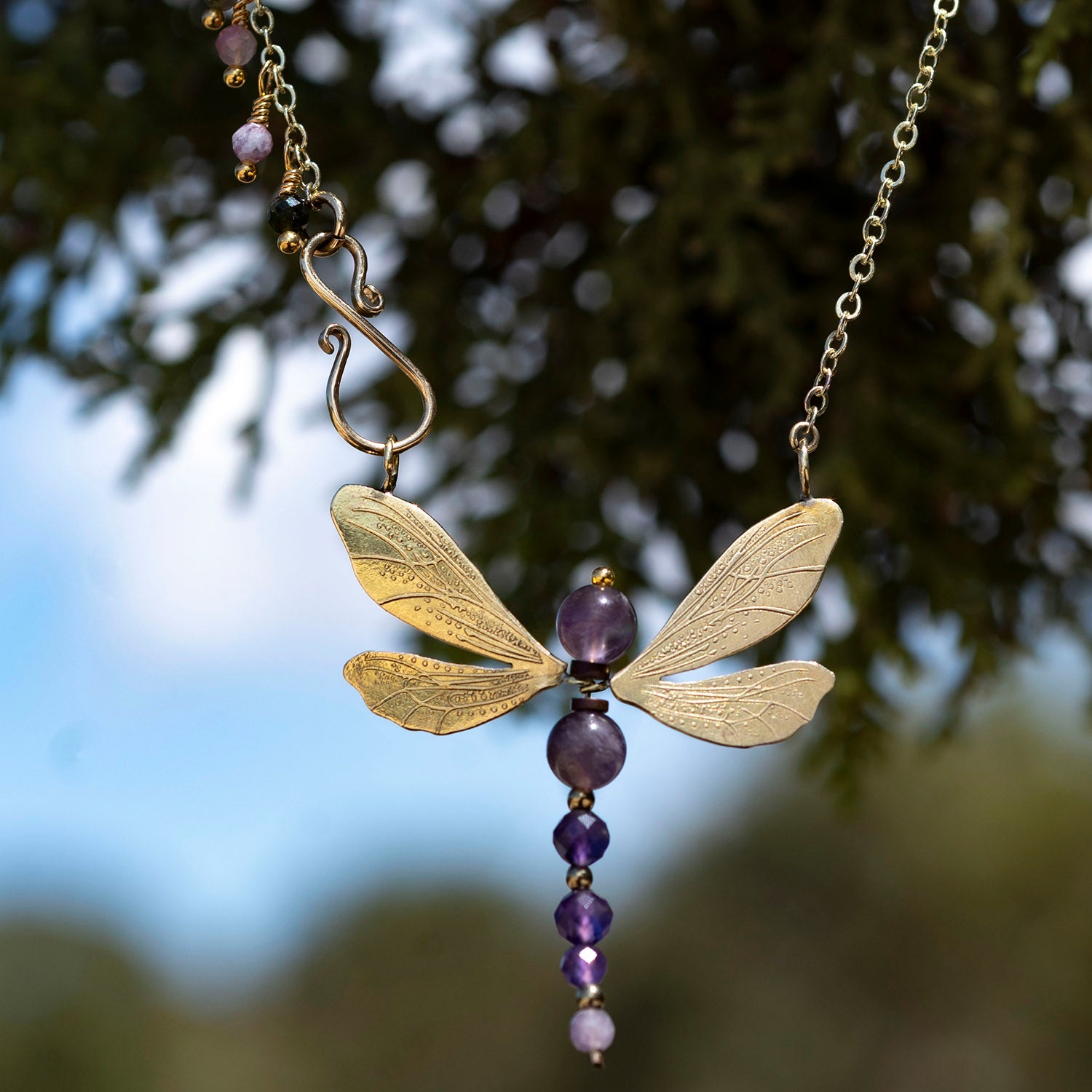 A dragonfly pendant necklace with textured gold wings and a beaded body made of purple and amethyst beads, hanging against a blurred background of green foliage and blue sky.