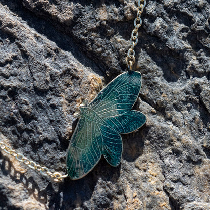 A gold-toned moth pendant necklace with a dark teal enamel fill, resting on a textured dark gray rock surface.
