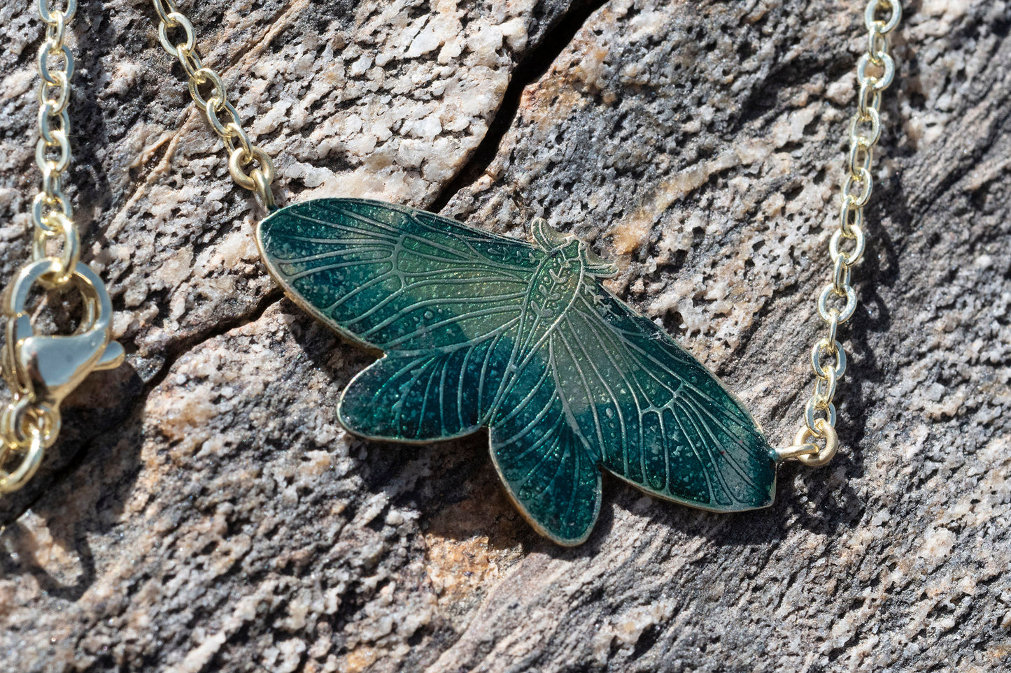 A close-up of a gold-tone chain necklace with a moth-shaped pendant featuring dark green and teal enamel, resting on a textured gray bark surface.
