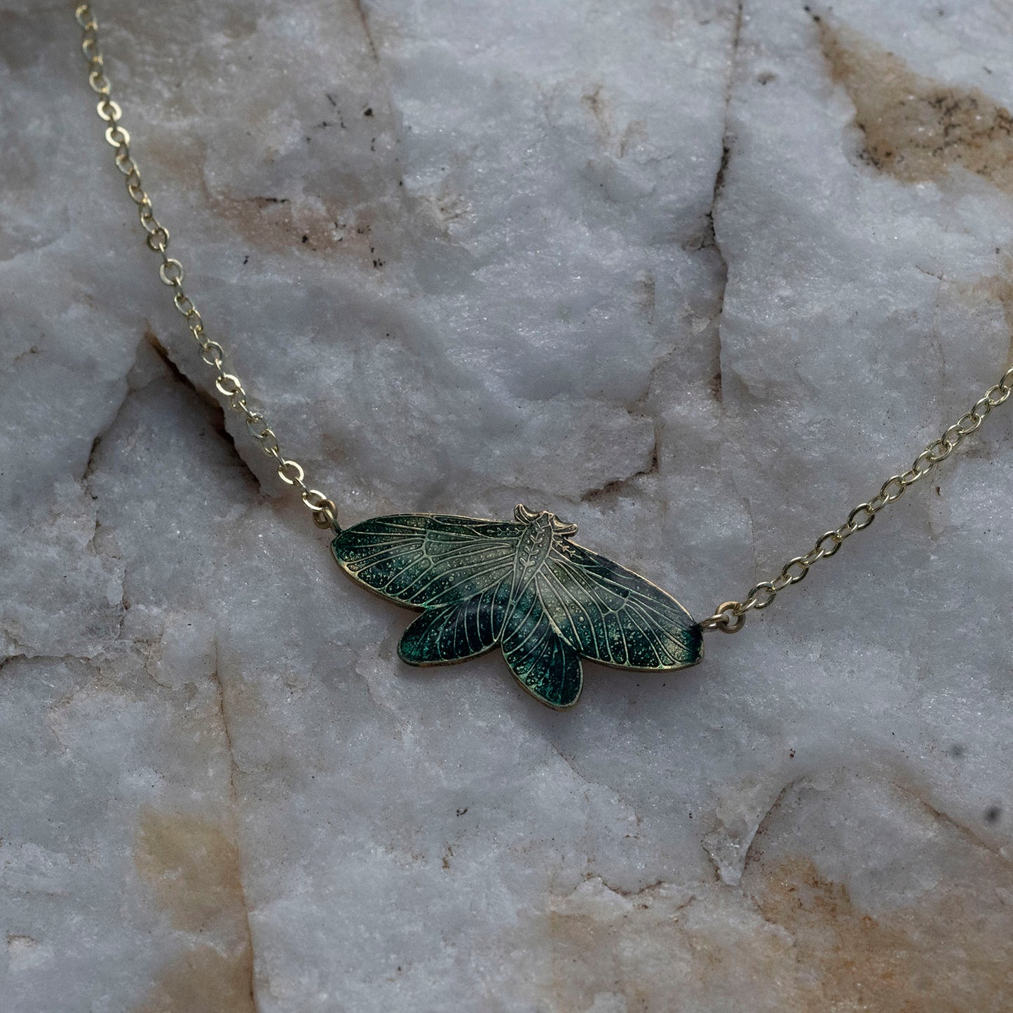 A delicate gold chain necklace featuring an intricately detailed moth pendant with dark green and gold enamel wings, resting on a textured white and gray stone surface.