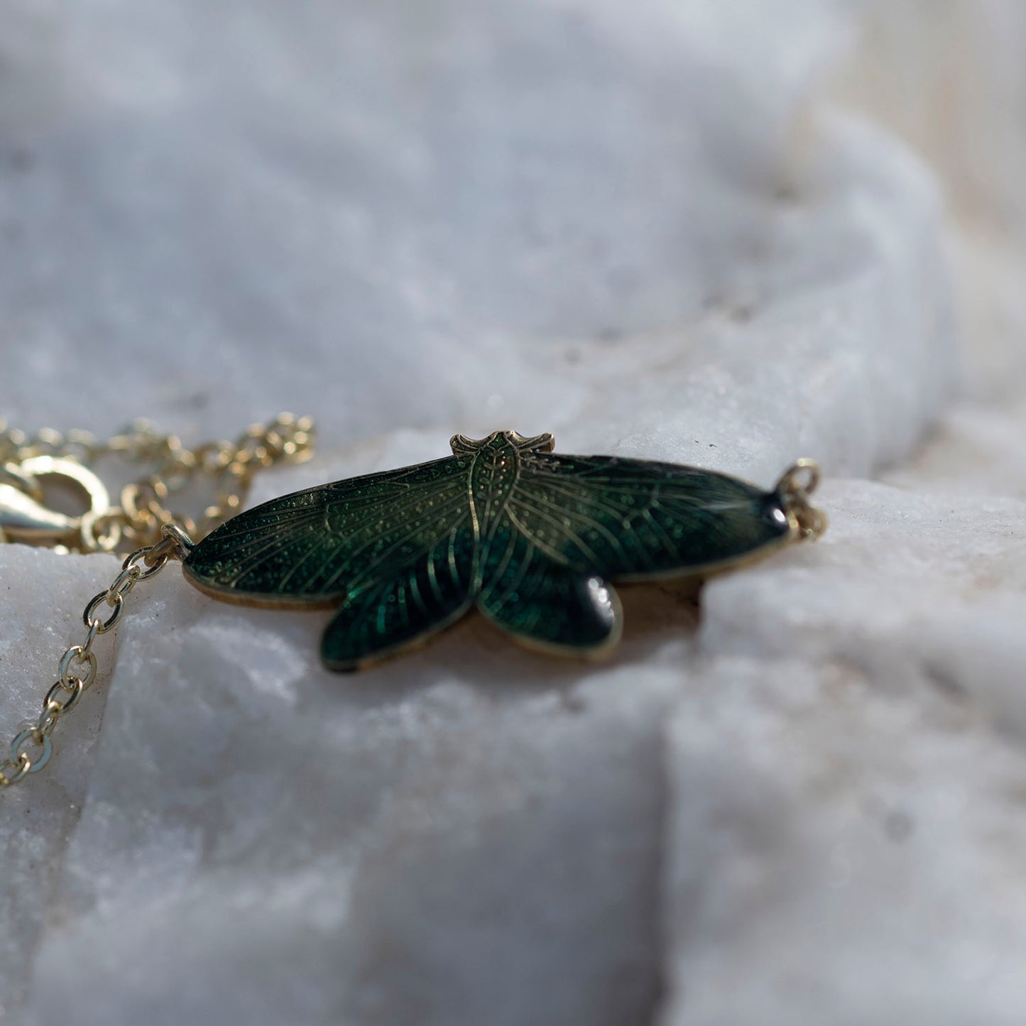 A close-up of a gold-tone pendant necklace shaped like a moth with dark green enamel wings, resting on a textured white stone surface.