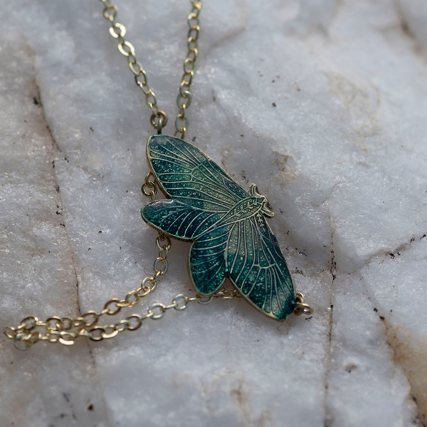 A close-up of a gold-tone chain necklace with a detailed moth pendant featuring dark green and teal enamel, resting on a textured white marble surface.