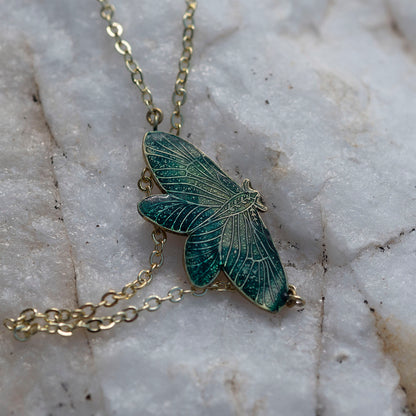 A close-up of a gold-tone chain necklace with a detailed moth pendant featuring dark green and teal enamel, resting on a textured white marble surface.