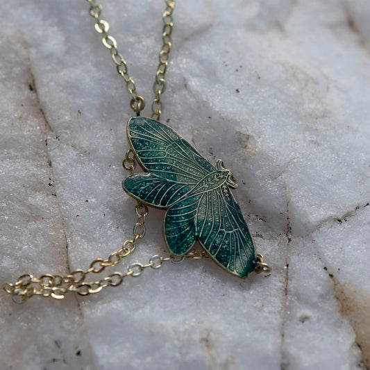 A close-up of a gold-tone chain necklace with a detailed moth pendant featuring dark green and teal enamel, resting on a textured white marble surface.