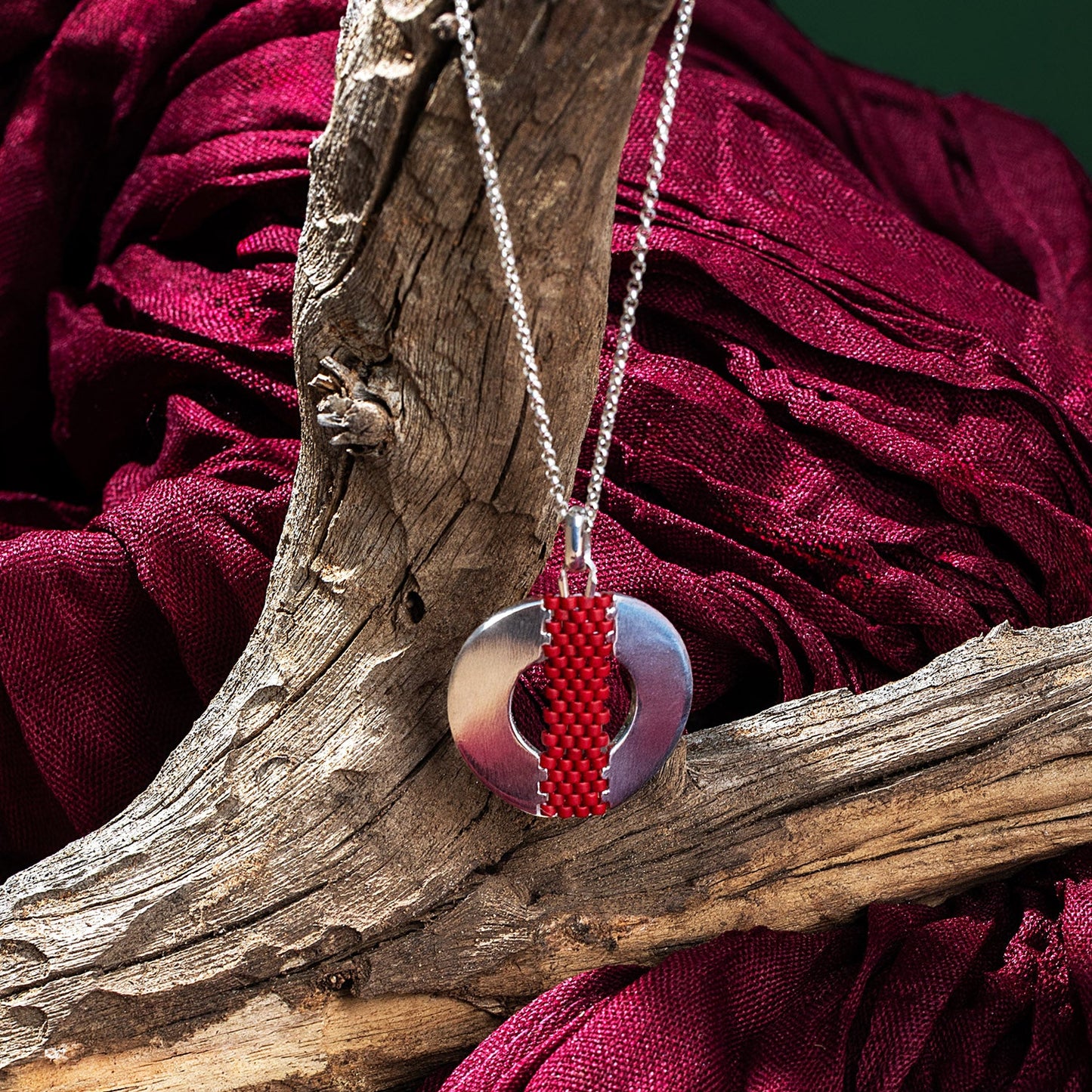 Necklace with a red and silver pendant on a wooden branch against a red fabric background