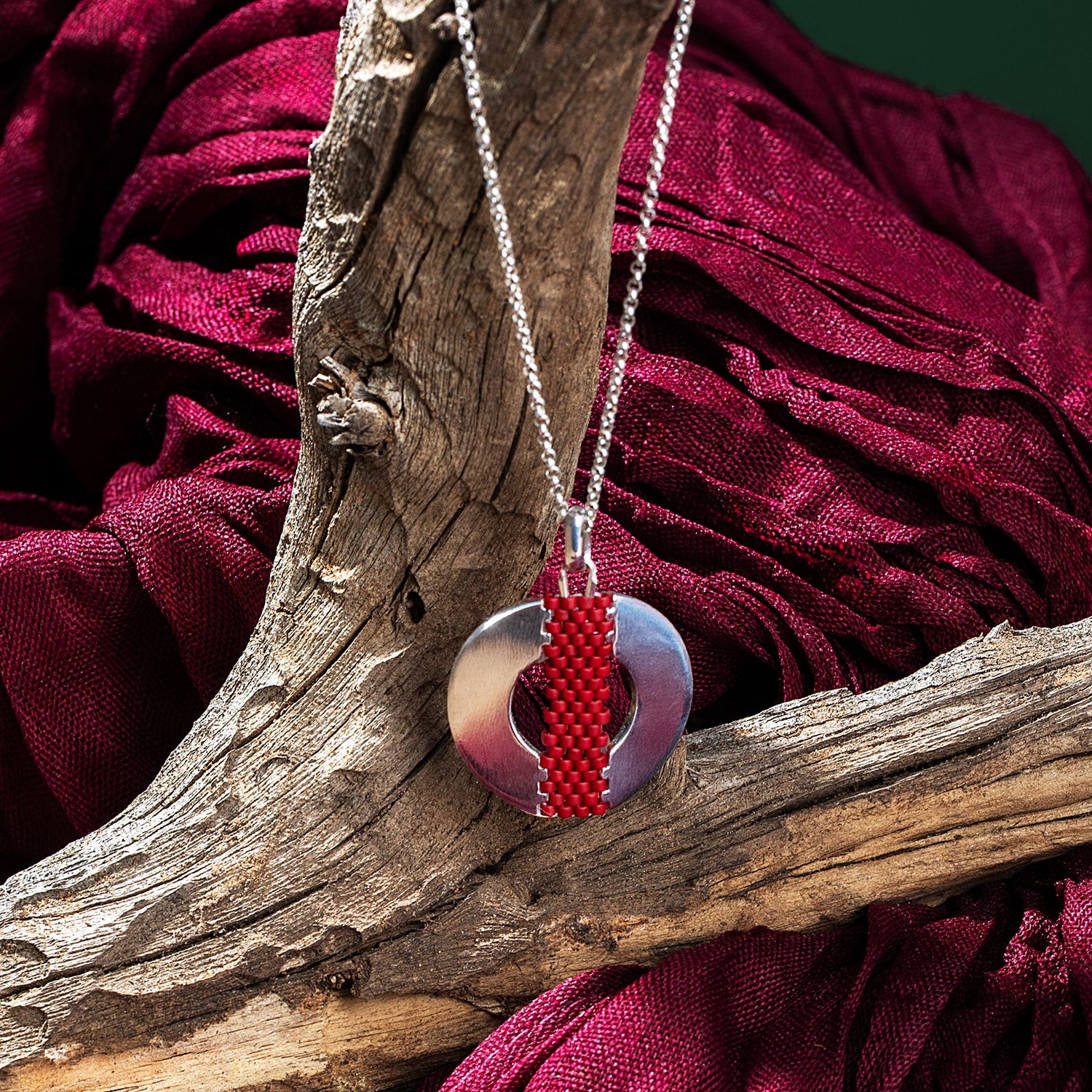 Necklace with a red and silver pendant on a wooden branch against a red fabric background