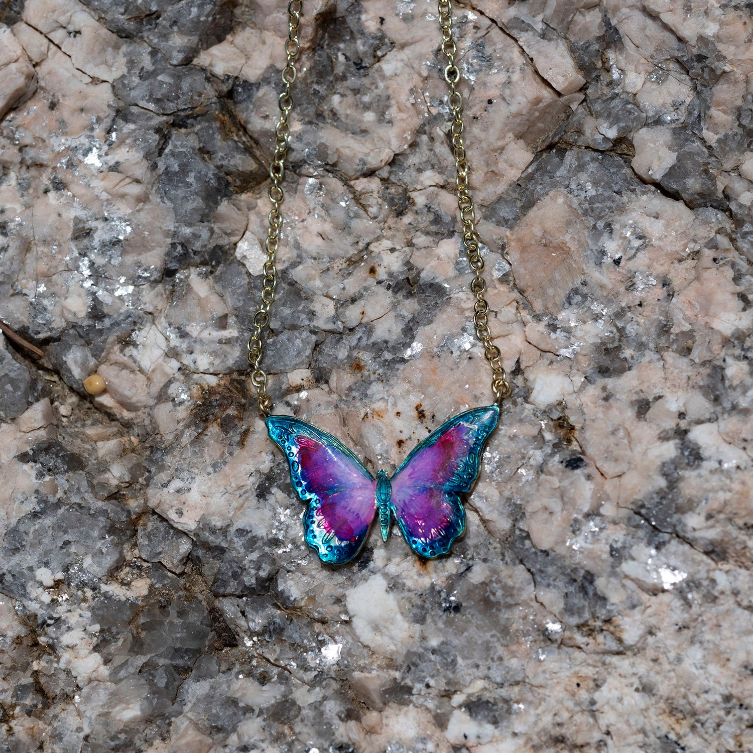 A butterfly pendant necklace with iridescent purple, pink, and blue wings on a gold chain, displayed on a textured gray and beige rock surface.