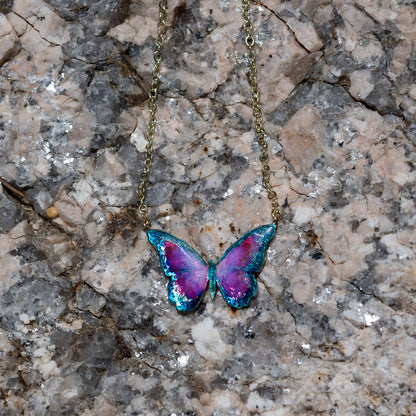 A butterfly pendant necklace with iridescent purple, pink, and blue wings on a gold chain, displayed on a textured gray and beige rock surface.