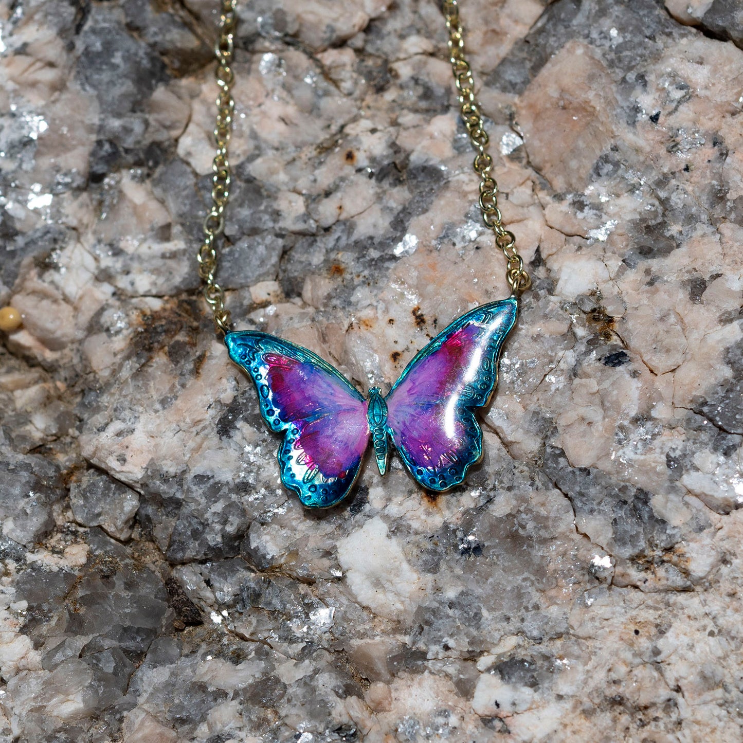 A butterfly-shaped pendant necklace with iridescent purple, blue, and teal enamel on a gold-tone chain, displayed on a textured gray and beige stone surface.
