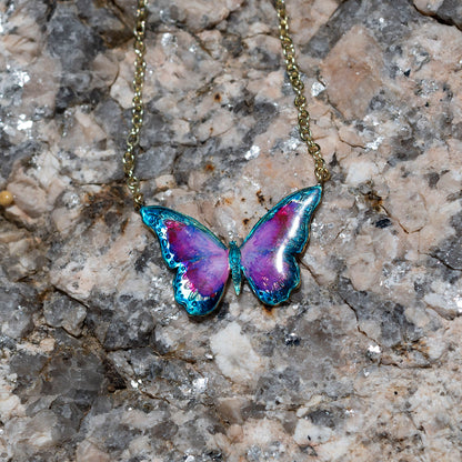 A butterfly-shaped pendant necklace with iridescent purple, blue, and teal enamel on a gold-tone chain, displayed on a textured gray and beige stone surface.