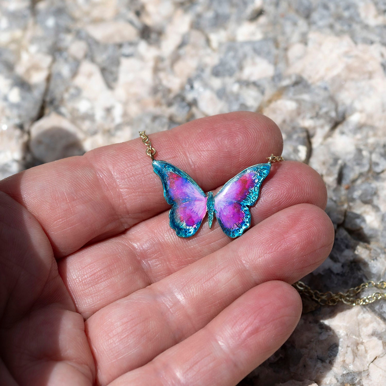 A close-up of a hand holding a small butterfly-shaped pendant necklace with iridescent pink, purple, and blue wings on a gold chain, set against a blurred rocky background.