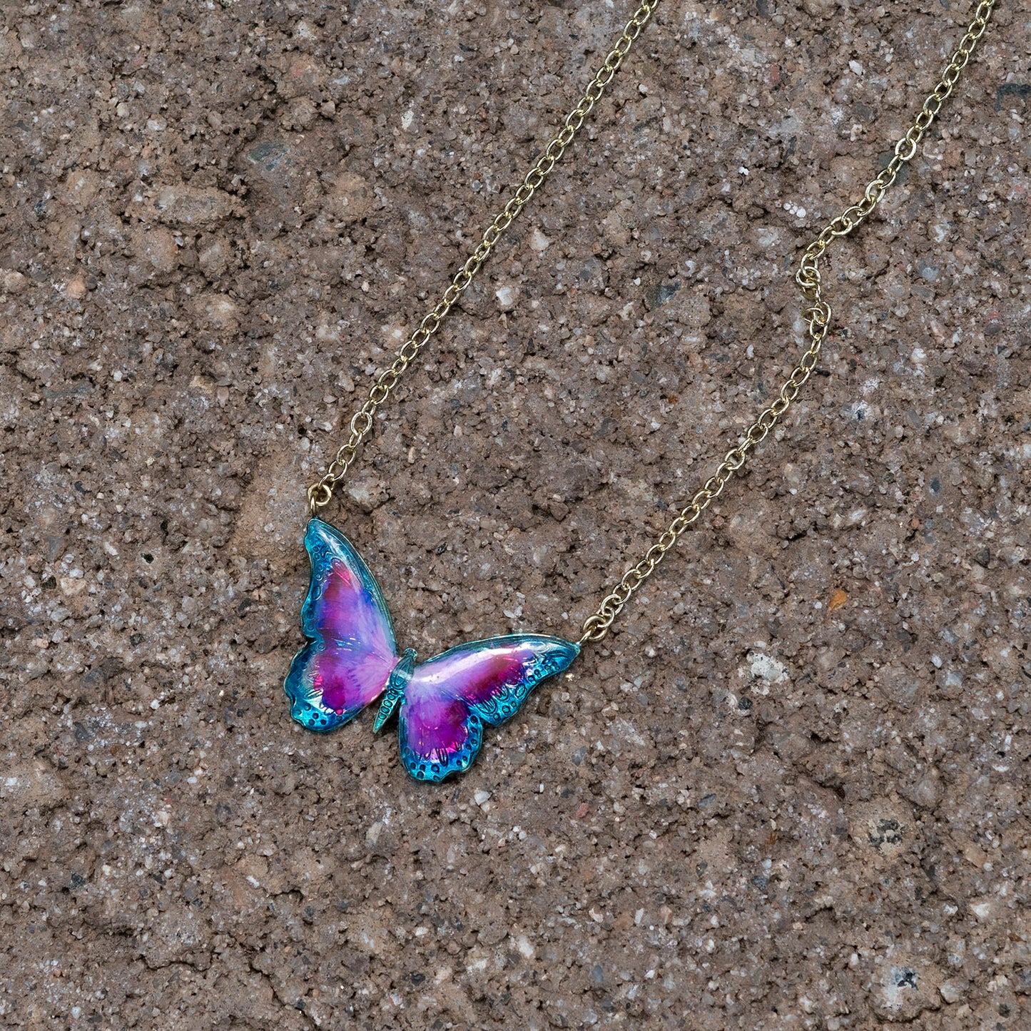 A gold-tone chain necklace with a butterfly pendant featuring vibrant purple and pink wings with teal-blue edges, displayed on a textured brown stone surface.