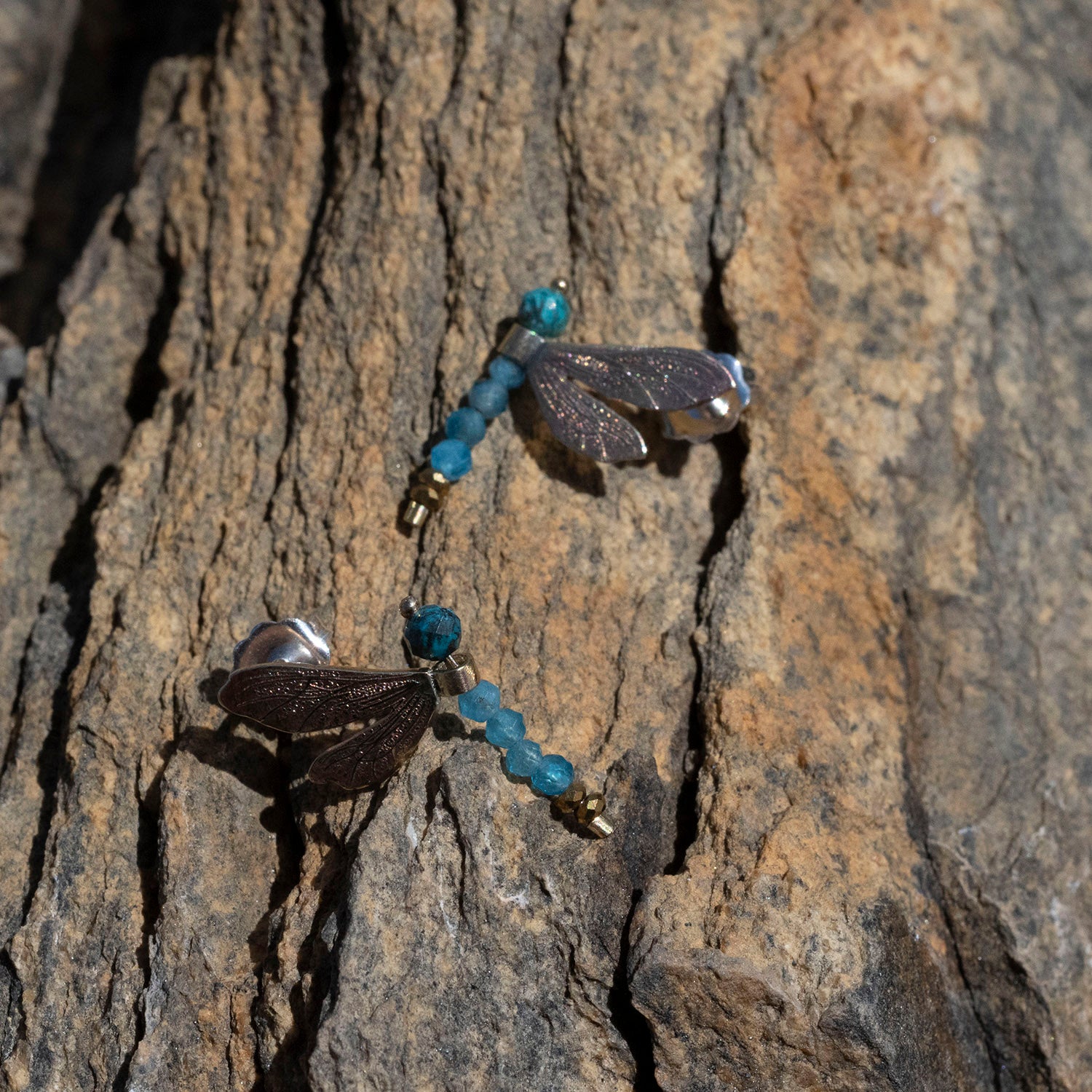 A pair of dragonfly earrings with dark brown wings, silver bodies, and blue-green beads, shown resting on a textured rock surface.