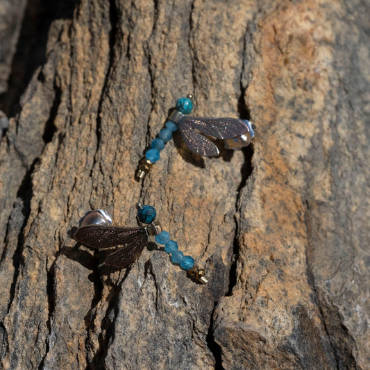 A pair of dragonfly earrings with dark brown wings, silver bodies, and blue-green beads, shown resting on a textured rock surface.
