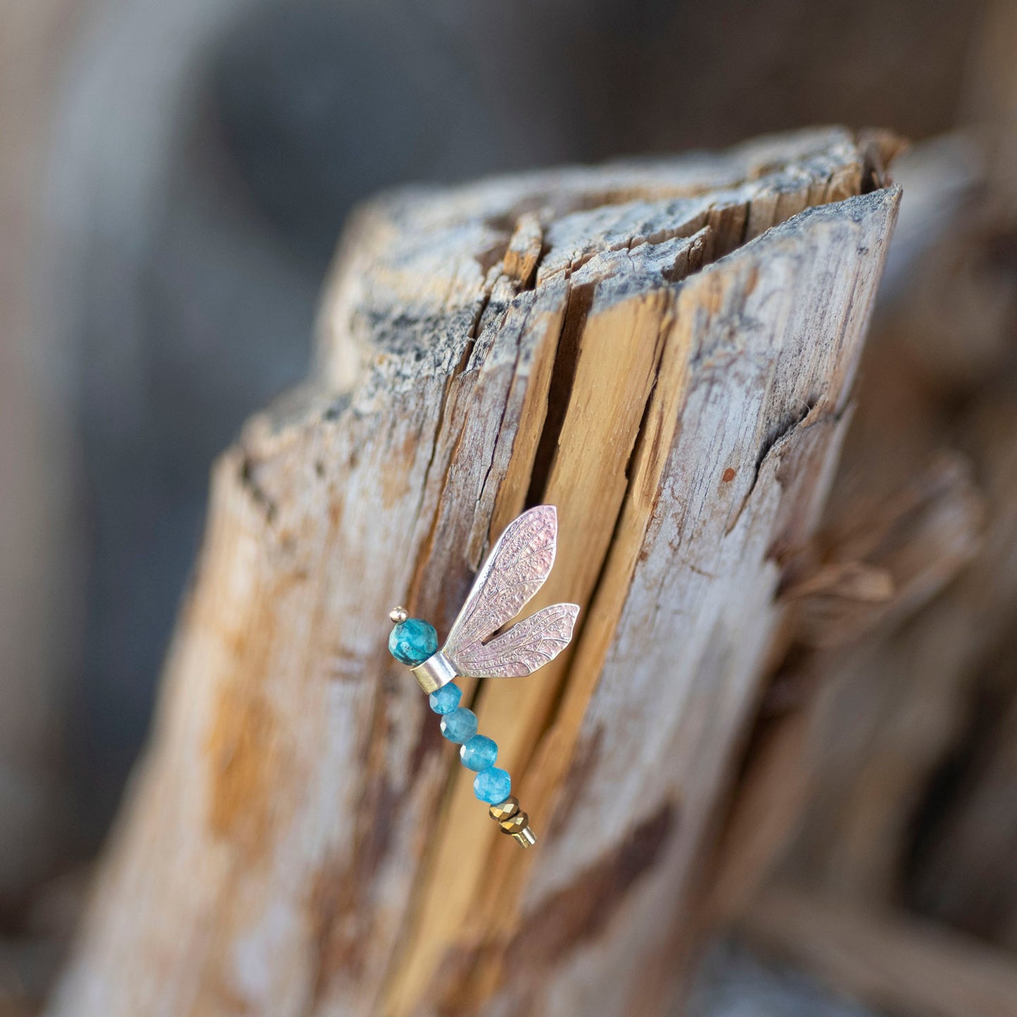A dragonfly-shaped earring with a textured rose gold body and translucent wings, accented with blue beads and gold spacer beads, resting on a piece of weathered driftwood.