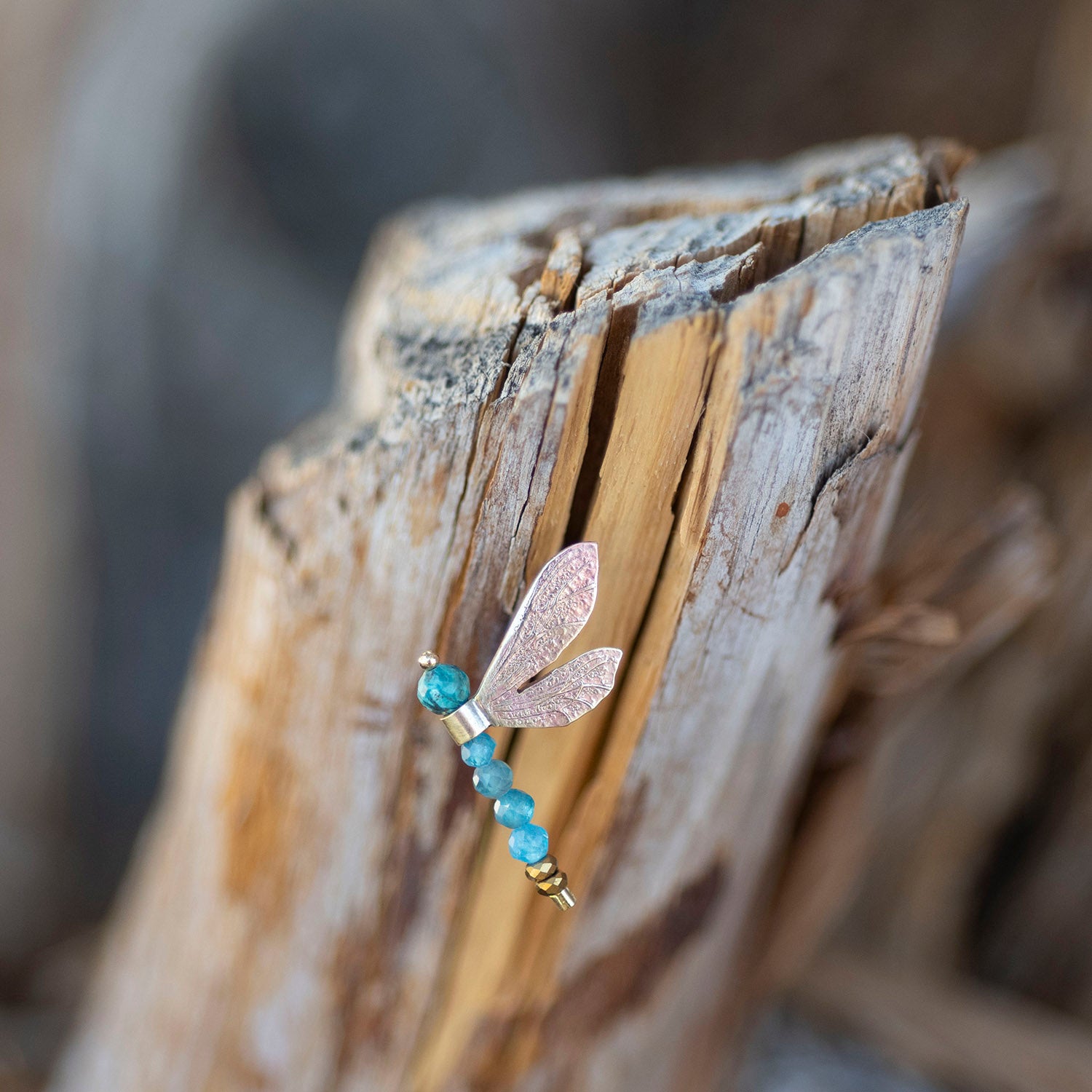 A dragonfly-shaped earring with a textured rose gold body and translucent wings, accented with blue beads and gold spacer beads, resting on a piece of weathered driftwood.