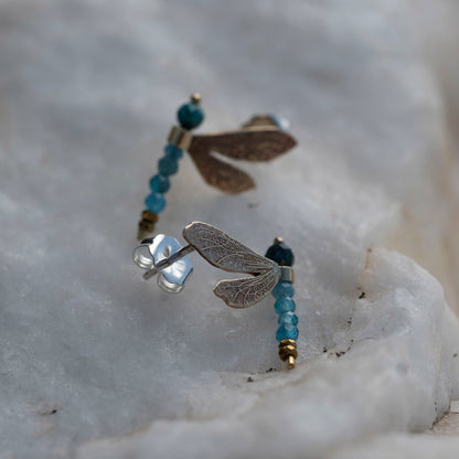 A pair of dragonfly stud earrings resting on a white crystalline stone surface. The earrings feature textured metallic wings (one silver-toned, one bronze-toned) and are accented with small blue beads and gold spacer beads.