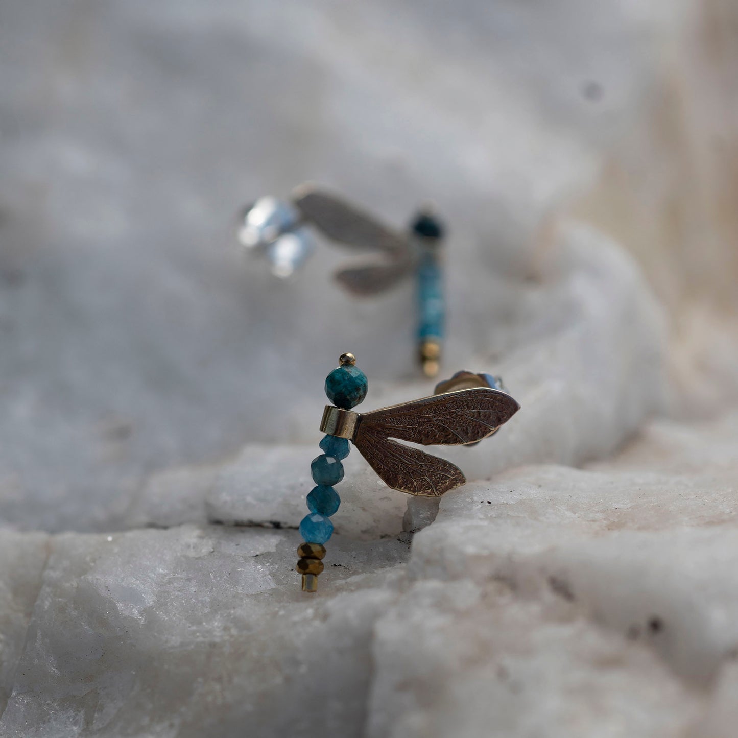 A pair of dragonfly earrings resting on a textured white stone surface. The front earring is in sharp focus, featuring a brown wing, a gold post, and a vertical bead strand with turquoise blue beads and a gold spacer.